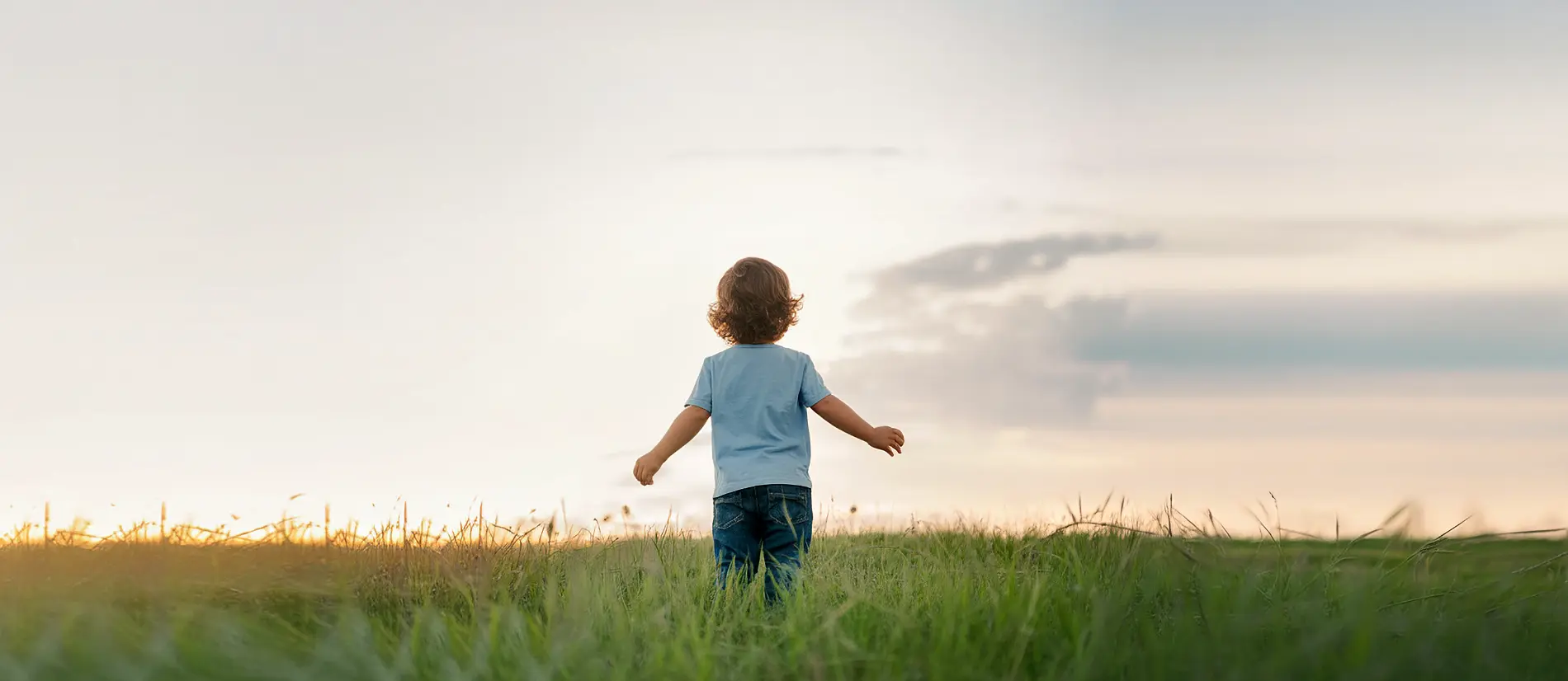 Kind steht mit dem Rücken zur Kamera in einem weiten Feld unter hellem, ruhigem Himmel. Die Arme sind leicht ausgebreitet, die Szene wirkt friedlich und hoffnungsvoll.
